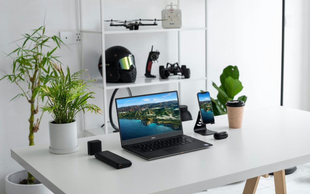 Mesa de escritório minimalista com notebook, smartphone, plantas decorativas e itens tecnológicos ao fundo.